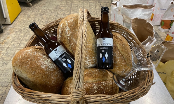 Twee flesjes Zwarte Parochie bier in een broodmand met vier broden en een leeg bierglas erin